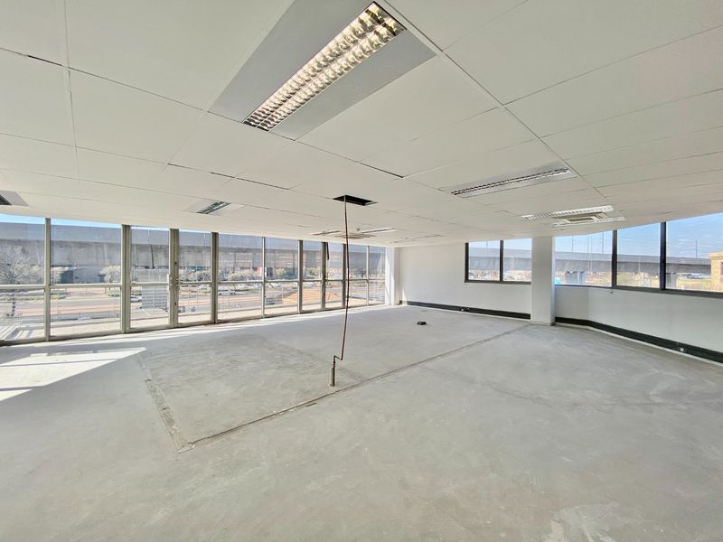 A vacant, open-plan commercial office space featuring floor-to-ceiling windows, suspended ceilings with recessed lighting, and unfinished concrete flooring.