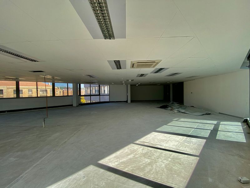A large, vacant open-plan commercial office space featuring suspended ceilings, recessed lighting, and expansive windows with natural light.