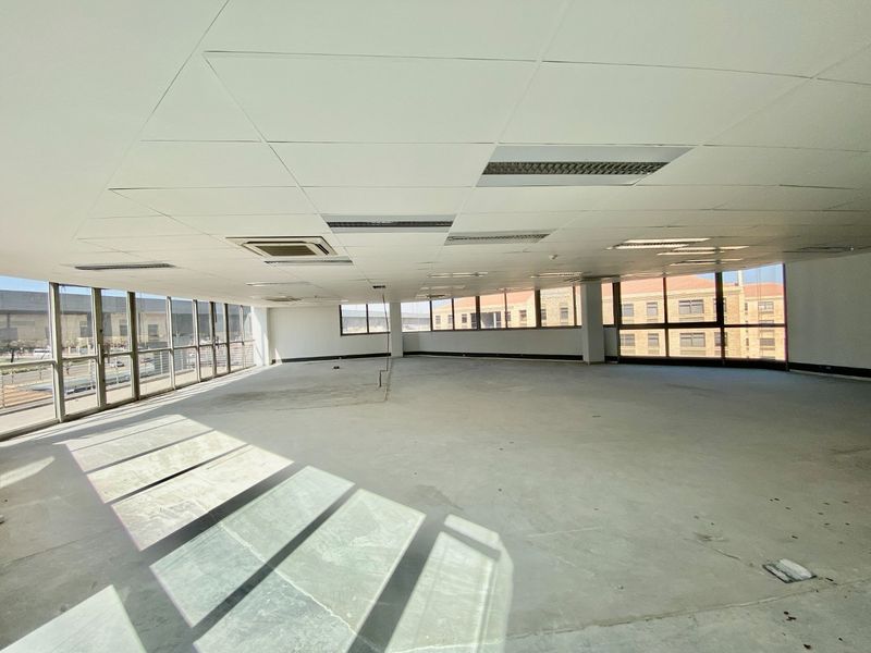 A spacious, vacant open-plan commercial office area featuring large windows, suspended ceilings with recessed lighting, and unfinished concrete flooring.