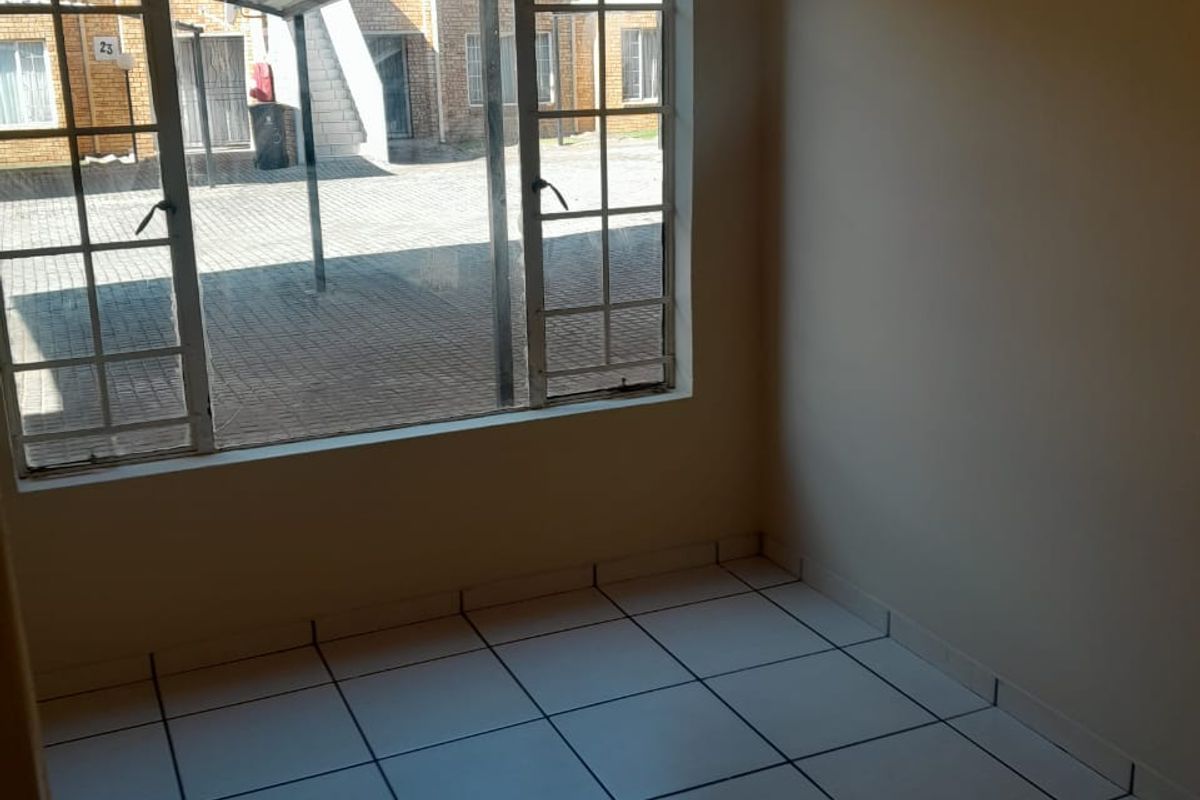 An empty, well-lit living area featuring clean tiled flooring and a large window overlooking the exterior complex area.