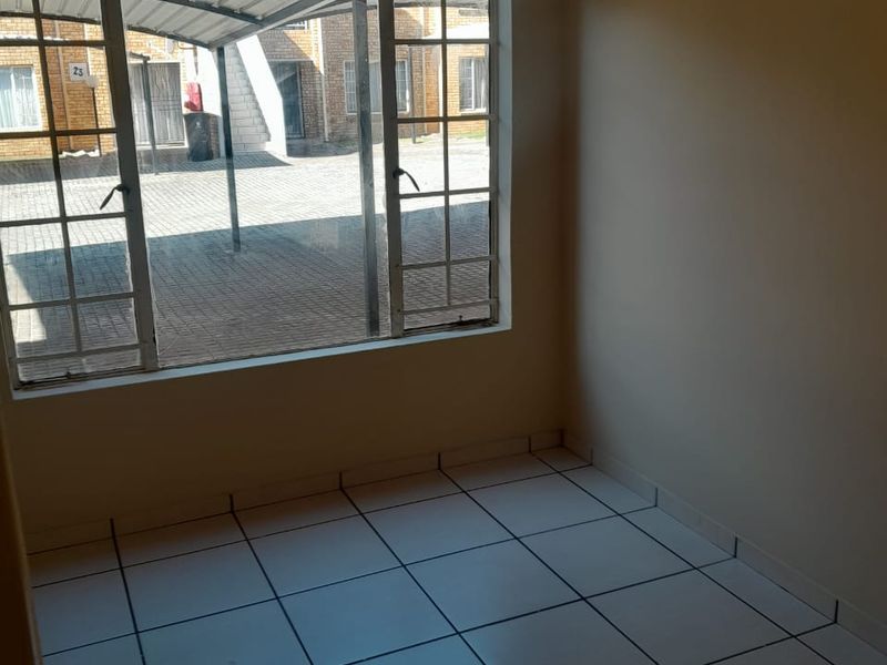 An empty, well-lit living area featuring clean tiled flooring and a large window overlooking the exterior complex area.