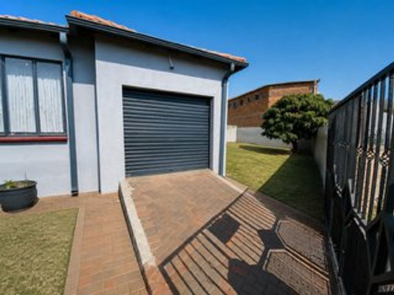Exterior view of a single garage with a roller door, paved driveway, and adjacent garden area, secured by a metal gate.