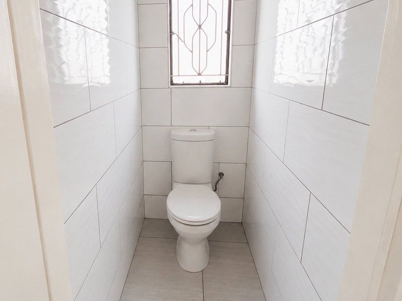 A clean, minimalist guest bathroom featuring modern white wall tiles, neutral floor tiles, and a secure window.