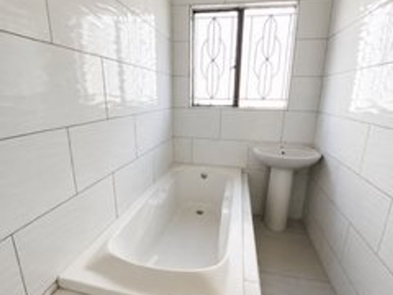 A clean, minimalist bathroom featuring a built-in bathtub, pedestal basin, and full-height wall tiling with a secure window.