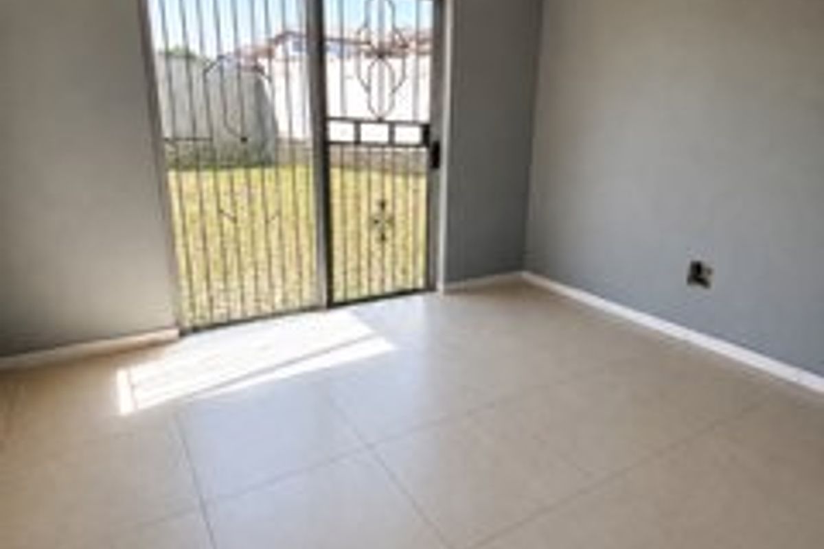 An empty living room featuring neutral tiled flooring, grey walls, and a sliding glass door with a security gate leading to the garden.
