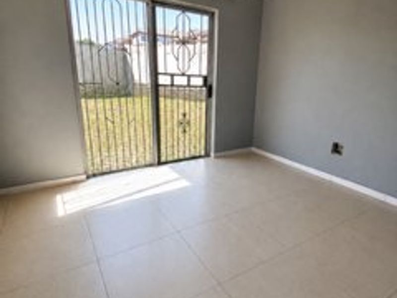 An empty living room featuring neutral tiled flooring, grey walls, and a sliding glass door with a security gate leading to the garden.