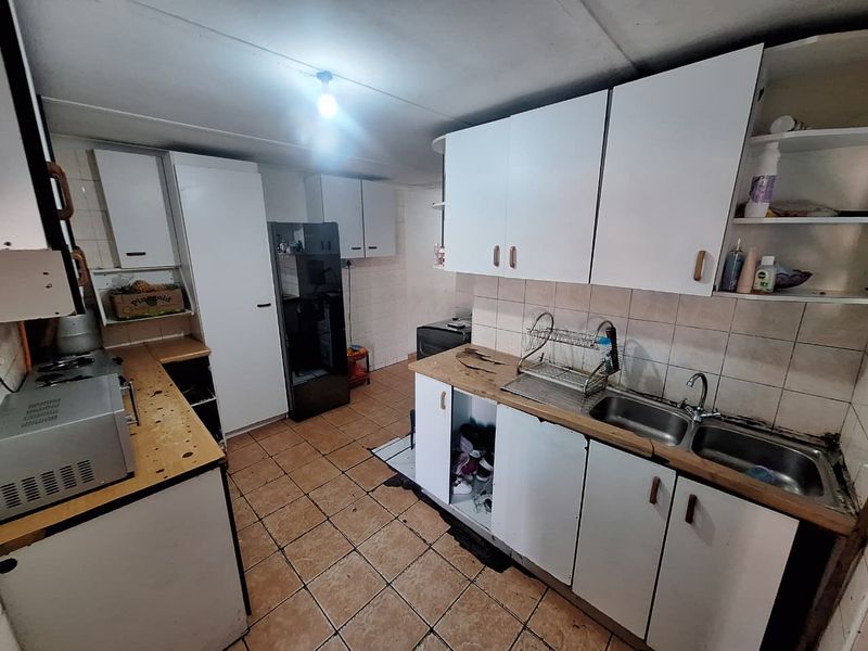 A functional kitchen featuring white cabinetry, double stainless steel sink, tiled splashbacks, and tiled flooring, currently requiring cosmetic updates and maintenance.