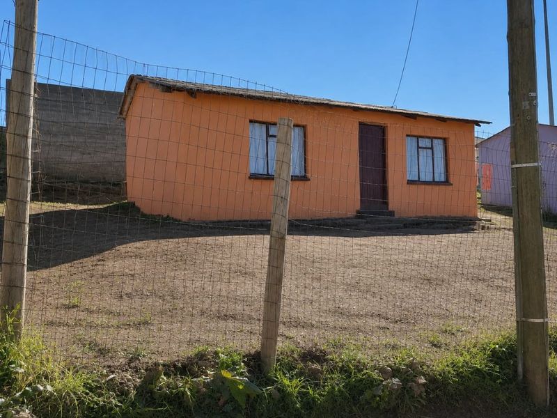 A modest single-story house with orange exterior walls, metal-framed windows, and a wire mesh perimeter fence on a dirt lot.
