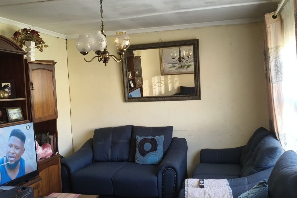 A living room featuring laminate flooring and a central chandelier, currently showing signs of wear on the ceiling and wall finishes.