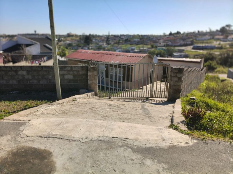 Front view of the property featuring a concrete driveway, metal entrance gate, and a concrete block boundary wall leading to the main dwelling.