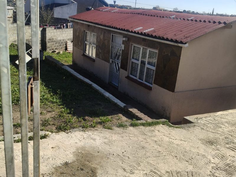 Front view of a modest single-story house featuring a corrugated metal roof, stone cladding accents, and a concrete driveway with a security gate.