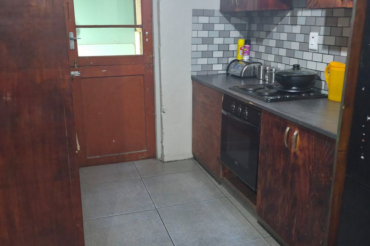 A compact kitchen featuring dark wood cabinetry, a tiled backsplash, built-in oven and hob, and tiled flooring, leading to an exterior timber door.
