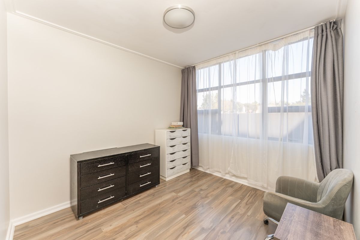A bright bedroom featuring laminate flooring, a large window with curtains, and a neutral color palette, offering a clean and functional living space.