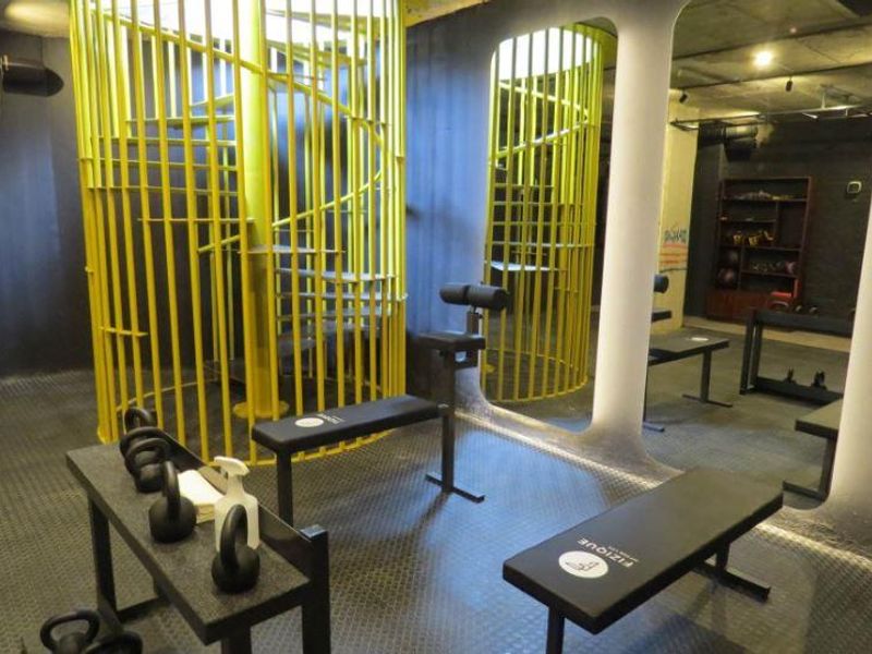 A modern gym space featuring industrial-style rubber flooring, a striking yellow spiral staircase, and a large mirrored wall with exposed concrete ceilings.