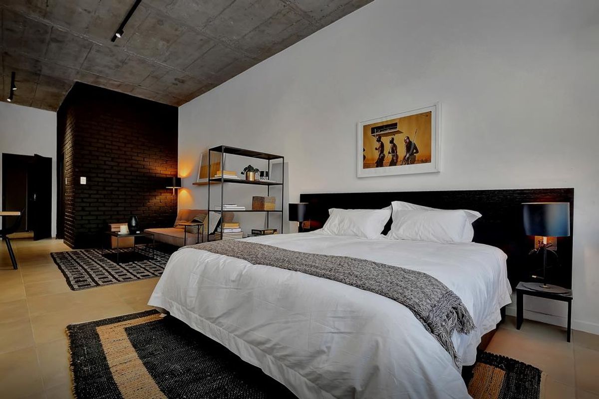 Spacious bedroom featuring an exposed concrete ceiling, a striking black brick feature wall, and modern tiled flooring with integrated track lighting.