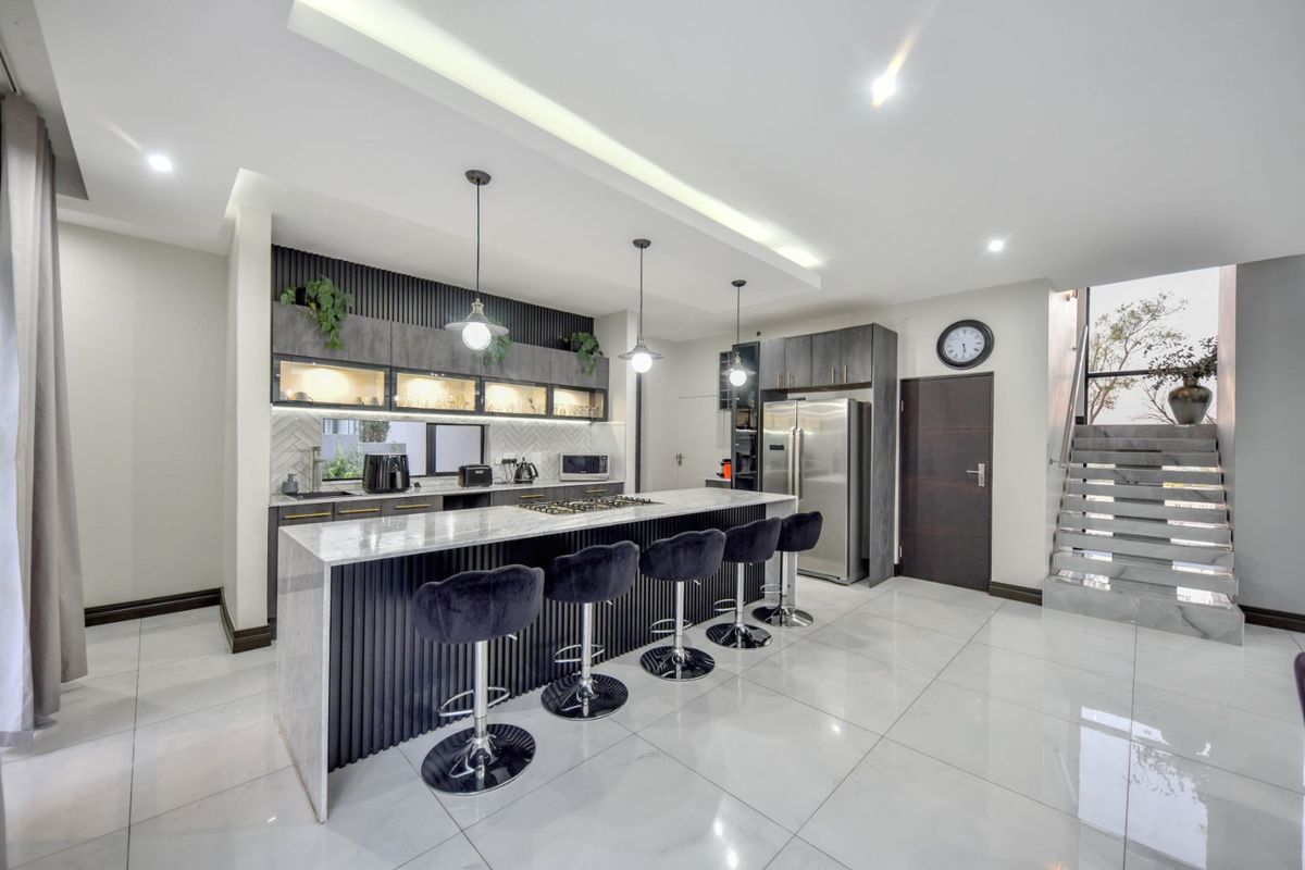Modern open-plan kitchen featuring a large stone-topped island, sleek dark cabinetry, integrated lighting, and high-gloss tiled flooring, adjacent to a contemporary staircase.