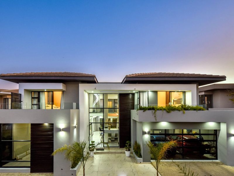 A modern, double-storey luxury residence featuring a striking glass entrance atrium, integrated garage, and contemporary architectural design with evening exterior lighting.