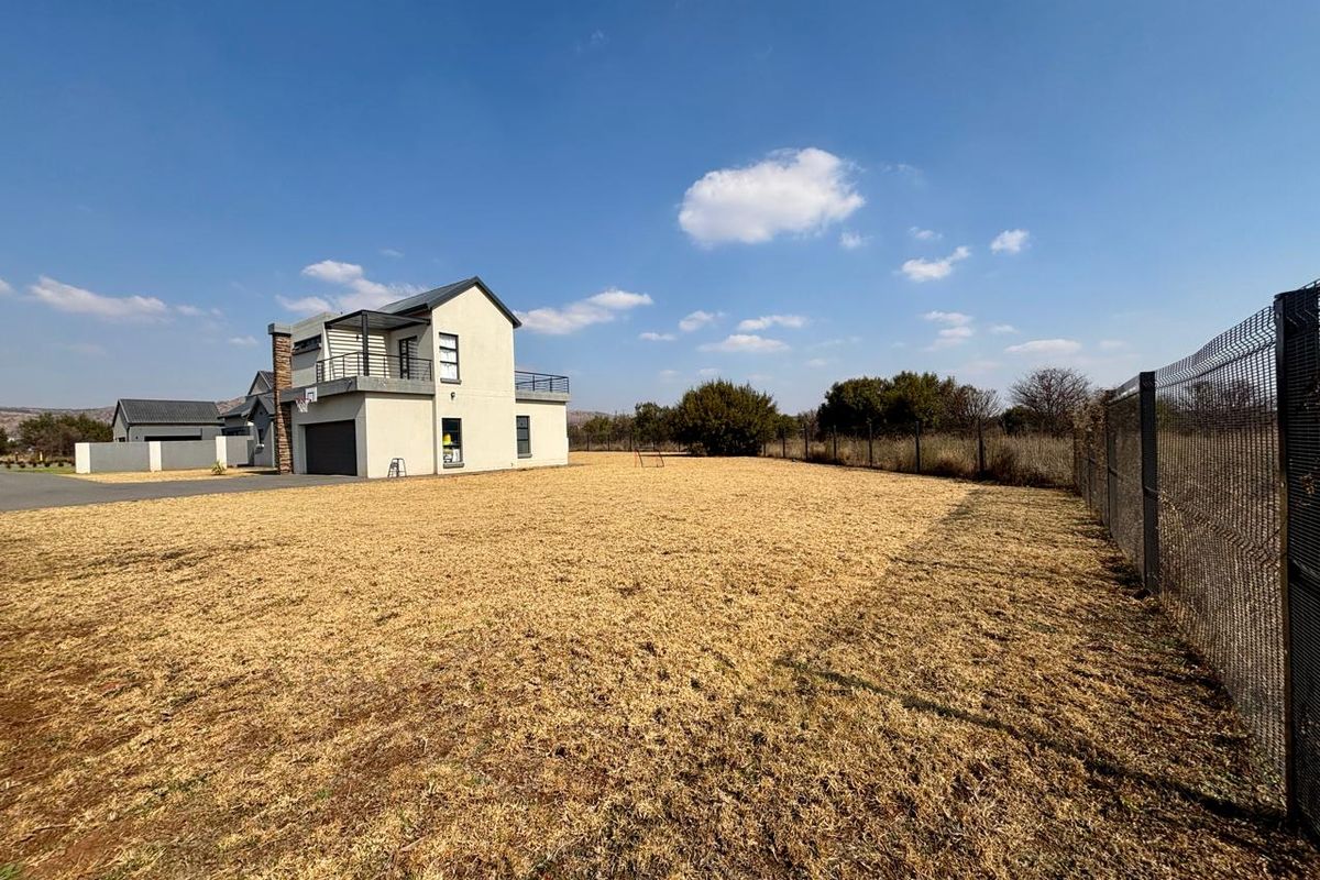 A spacious rear yard featuring a large lawn area, perimeter mesh fencing, and a view of the modern two-story house with a balcony and garage.