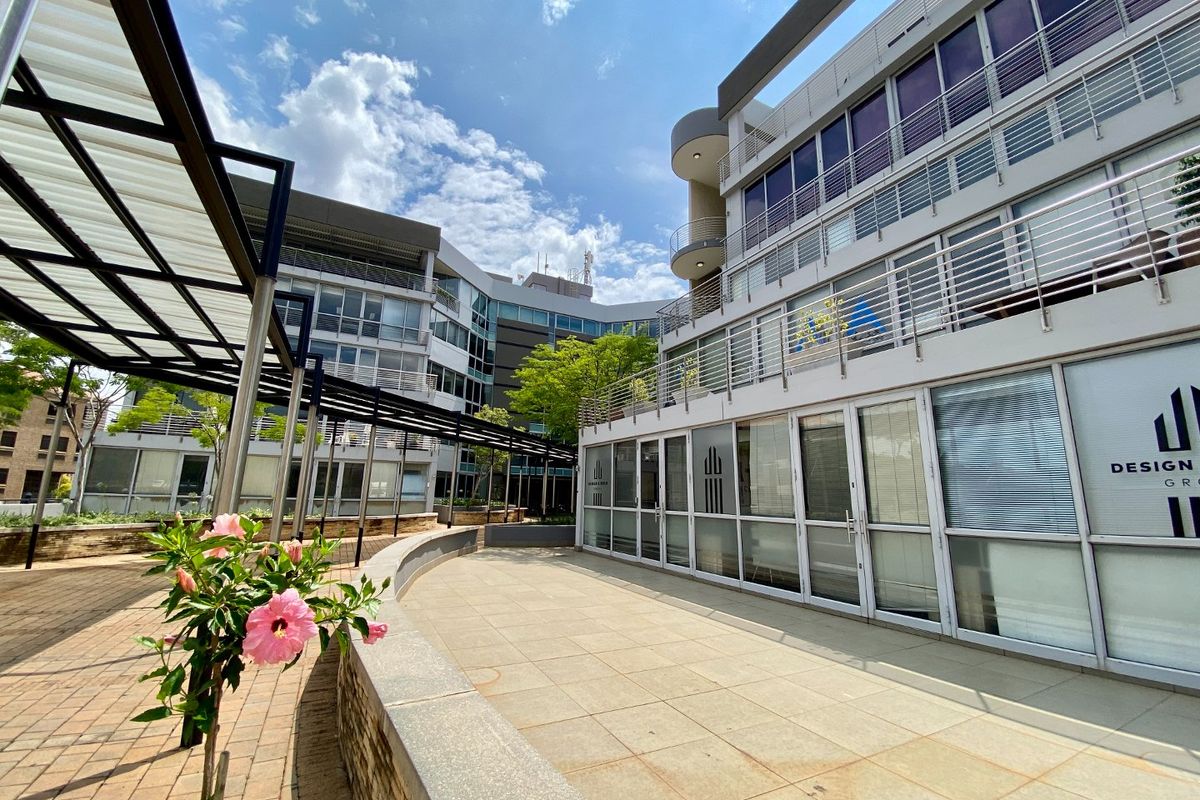 A modern commercial office complex featuring a paved courtyard, glass-fronted office units, and multi-level balconies with metal railings.