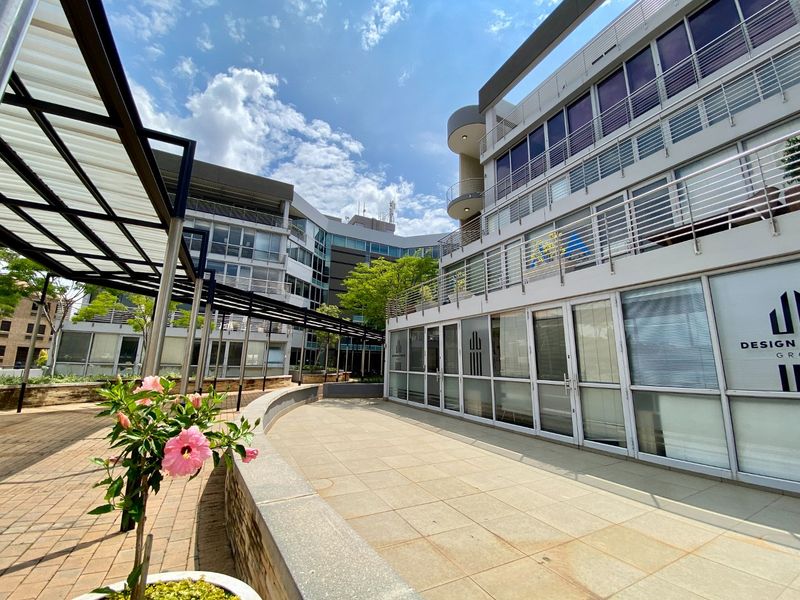 A modern commercial office complex featuring a paved courtyard, glass-fronted office units, and multi-level balconies with metal railings.