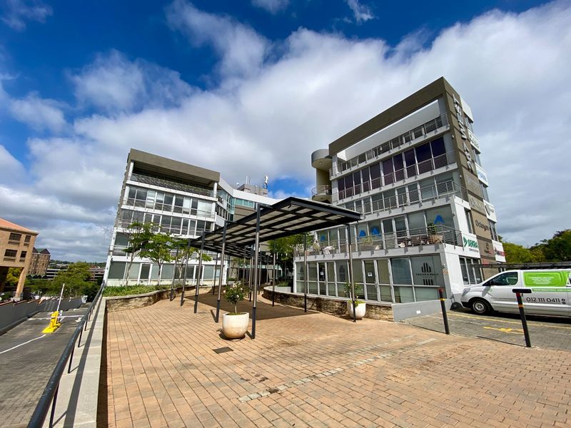 Modern multi-story commercial office building featuring a paved entrance courtyard with a covered walkway and glass facade elements.