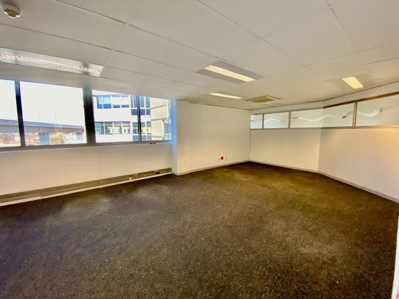 An empty commercial office space featuring large windows, suspended ceilings with recessed lighting, and carpeted flooring, suitable for a professional workspace.