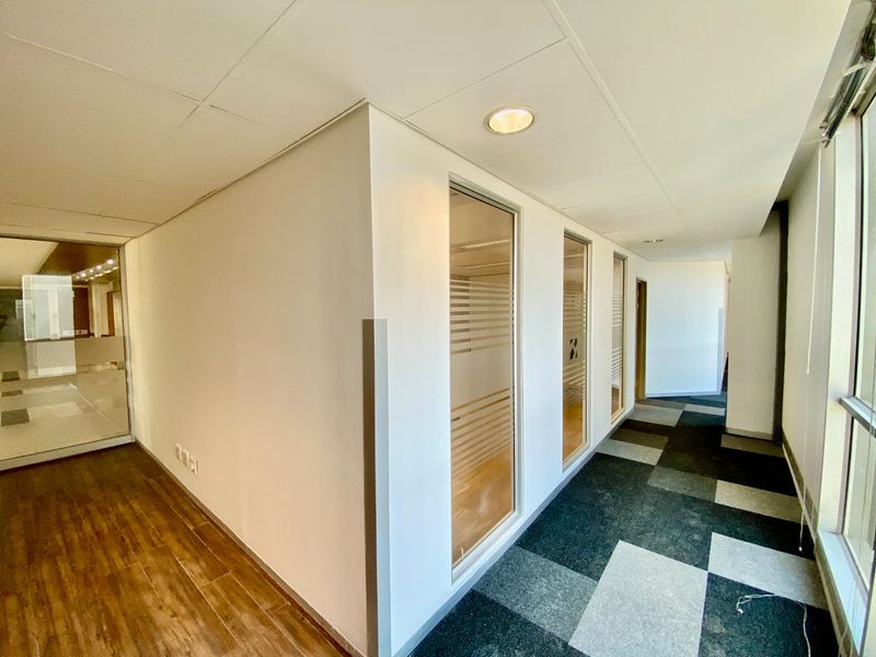 A bright, modern commercial hallway featuring a mix of wood-look and carpet tile flooring, with glass partitions providing visibility into adjacent office spaces.