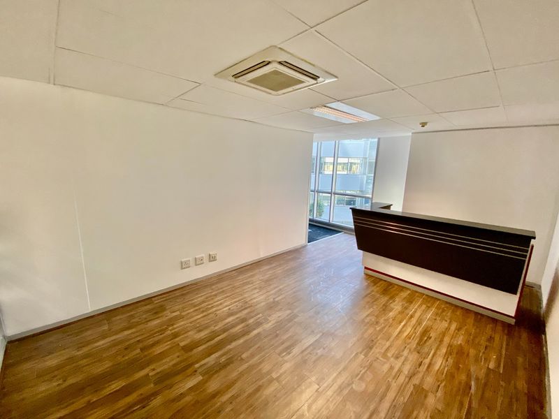 A bright, vacant commercial office space featuring wood-look flooring, a built-in reception desk, and a suspended ceiling with integrated air conditioning.