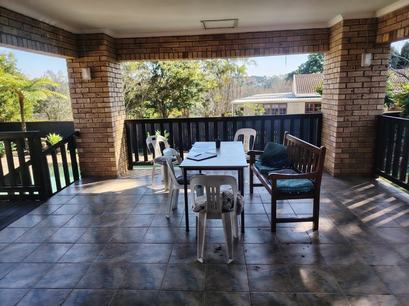 A covered outdoor patio featuring exposed brick pillars, tiled flooring, and a timber balustrade overlooking the garden and pool area.
