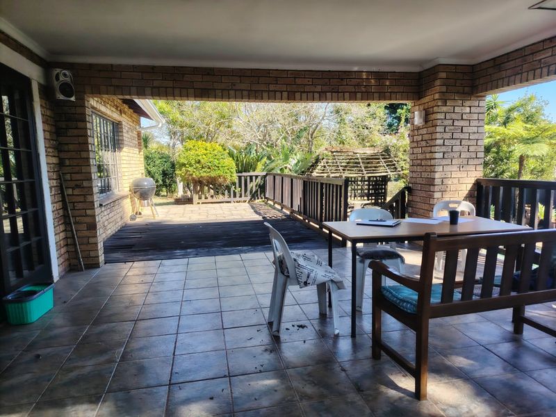 Covered outdoor patio featuring brick pillars and tiled flooring, extending onto a timber deck area overlooking the garden.