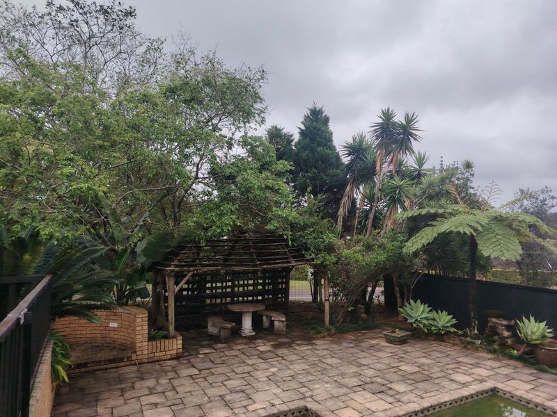 Outdoor pool area featuring a paved patio, brick retaining wall, and a weathered wooden pergola structure surrounded by established garden landscaping.