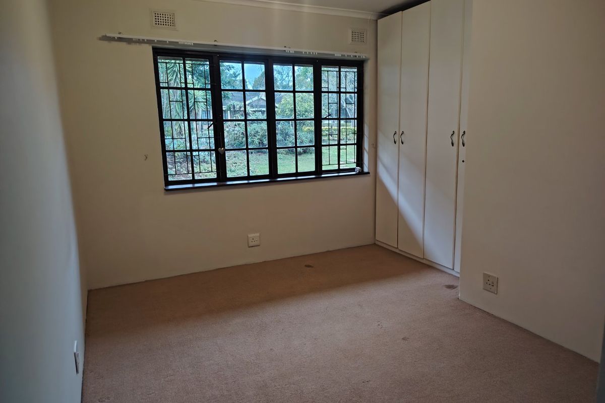 A vacant bedroom featuring built-in cupboards, carpeted flooring, and a window fitted with security bars overlooking the garden.