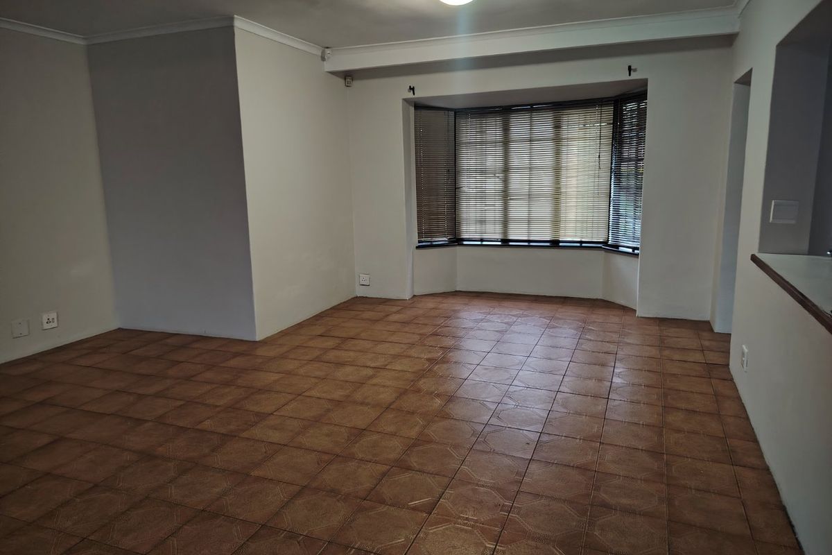 A spacious, empty living room featuring tiled flooring, a large bay window with fitted venetian blinds, and neutral wall finishes.