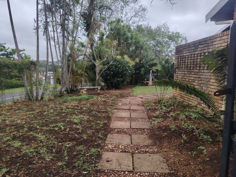 A garden area featuring a paved walkway alongside a brick boundary wall, surrounded by established trees and sparse lawn.