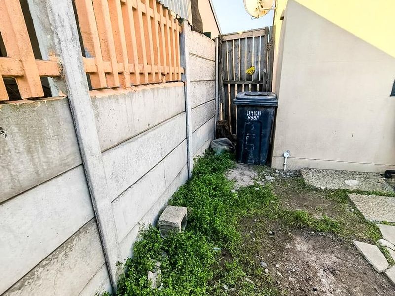 A narrow rear exterior side passage featuring a precast concrete boundary wall, a wooden gate, and an unpaved ground area with overgrown vegetation.
