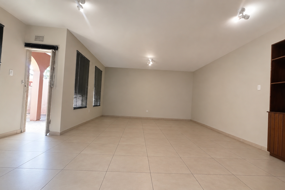 Spacious, light-filled living area featuring neutral tiled flooring, built-in wooden cabinetry, and large windows with venetian blinds, offering a clean and versatile space.