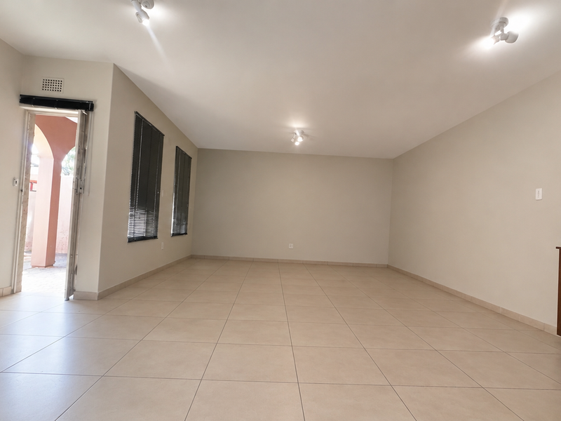 Spacious, light-filled living area featuring neutral tiled flooring, built-in wooden cabinetry, and large windows with venetian blinds, offering a clean and versatile space.