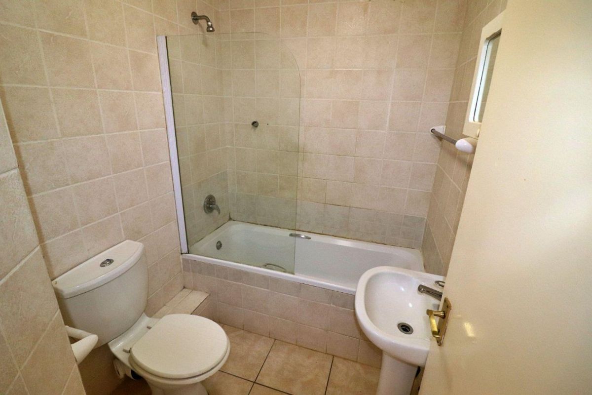 A functional bathroom featuring a pedestal basin, toilet, and a bathtub with a glass shower screen, finished with neutral wall and floor tiling.