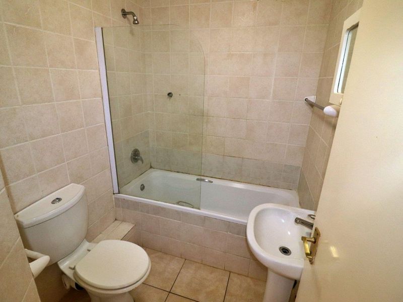 A functional bathroom featuring a pedestal basin, toilet, and a bathtub with a glass shower screen, finished with neutral wall and floor tiling.