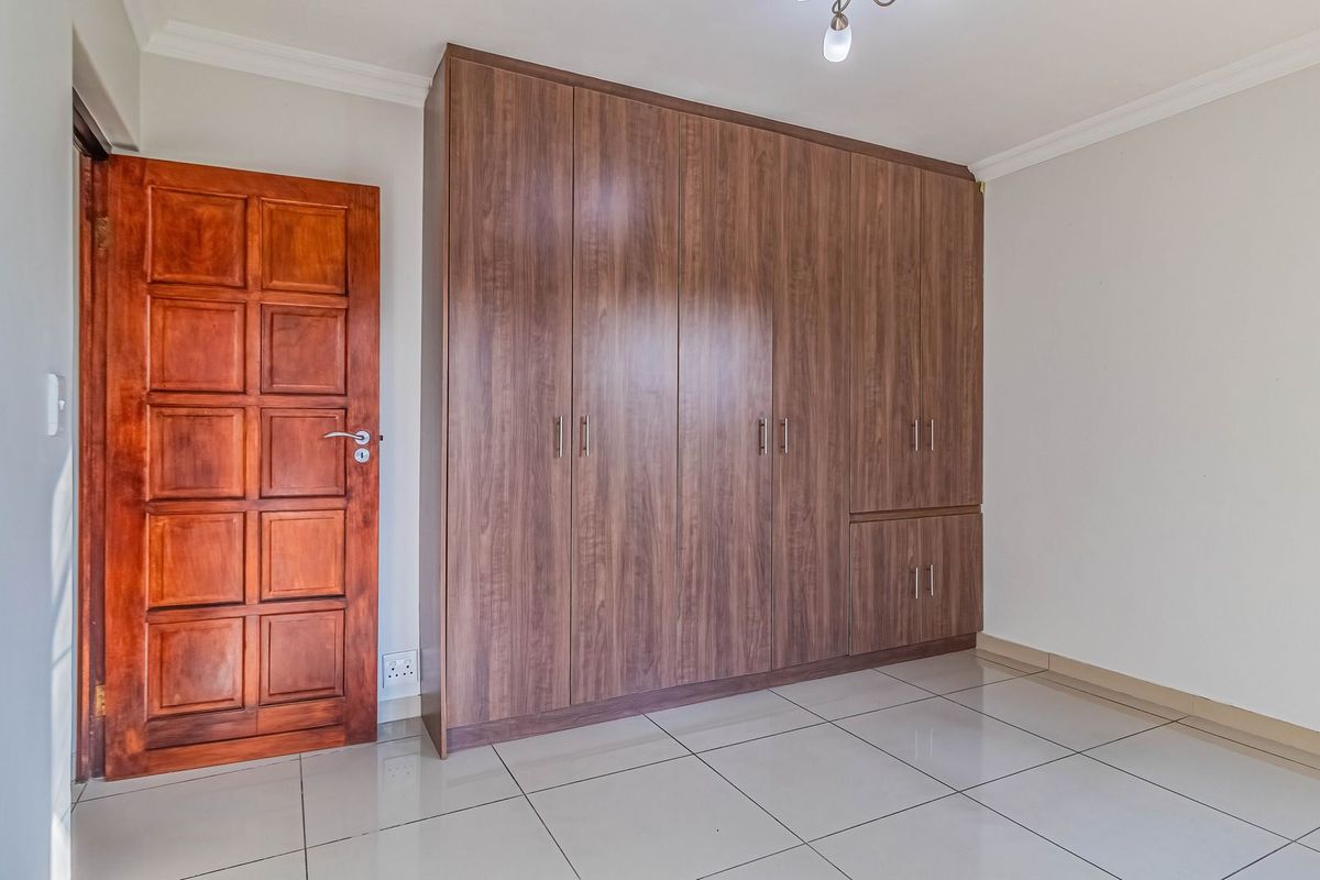 A clean, empty bedroom featuring modern wood-finish built-in cupboards, neutral tiled flooring, and a solid wooden door.