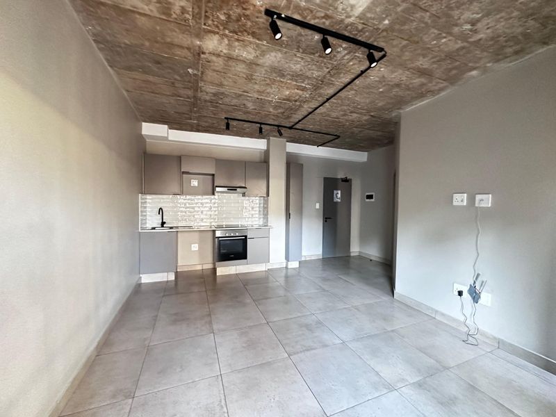 Modern open-plan living area featuring an exposed concrete ceiling, track lighting, and a compact kitchen with built-in appliances and tiled flooring.