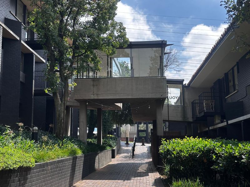 A view of the apartment complex exterior featuring a raised concrete walkway, brick paving, and landscaped garden areas between the building blocks.