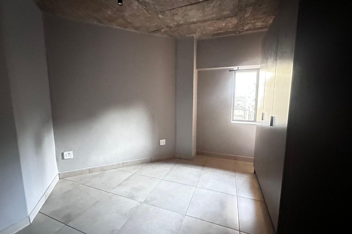 A minimalist bedroom featuring tiled flooring, built-in cupboards, and an industrial-style exposed concrete ceiling with natural light from a single window.