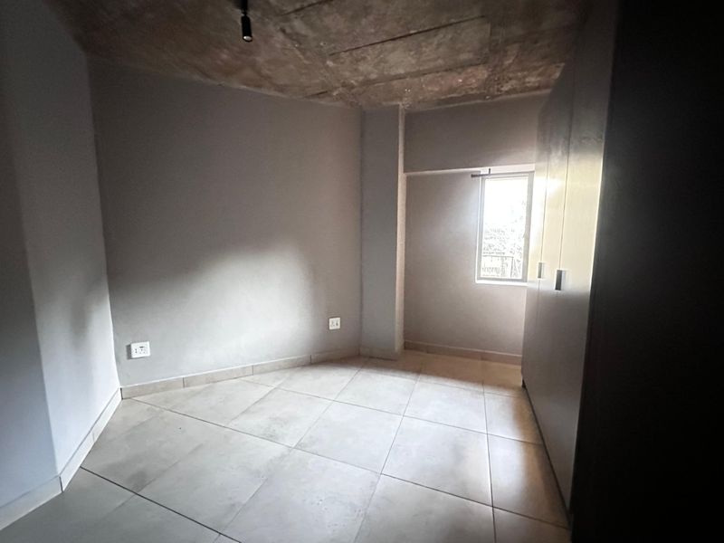 A minimalist bedroom featuring tiled flooring, built-in cupboards, and an industrial-style exposed concrete ceiling with natural light from a single window.