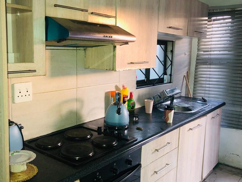 A compact kitchen featuring light wood-finish cabinetry, a built-in oven and hob, stainless steel sink, and tiled flooring, requiring some cosmetic updates.