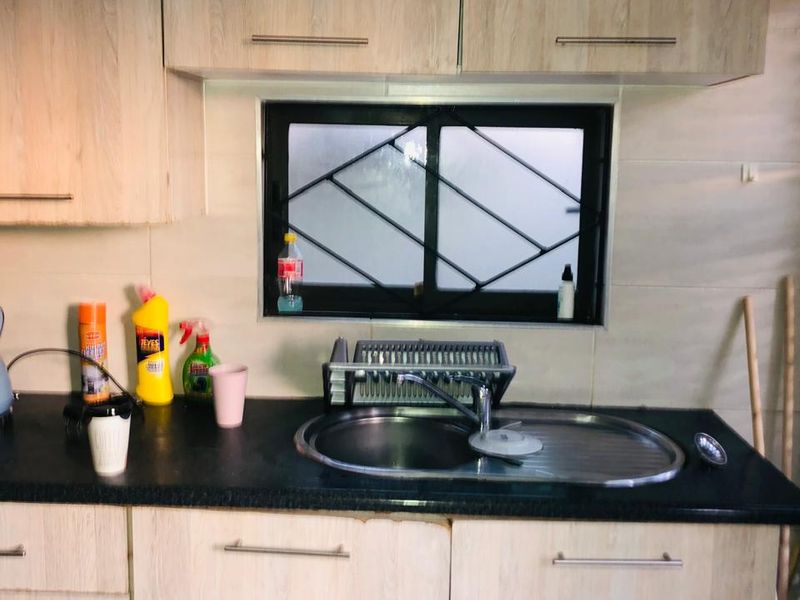 A functional kitchen area featuring light wood-look cabinetry, a black granite countertop, a stainless steel sink, and a window with security bars.