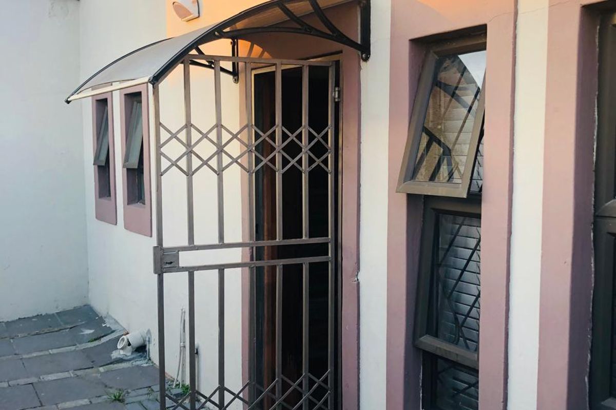 Rear exterior view featuring a security gate, protective awning, casement windows, and paved patio area with an external water tap.