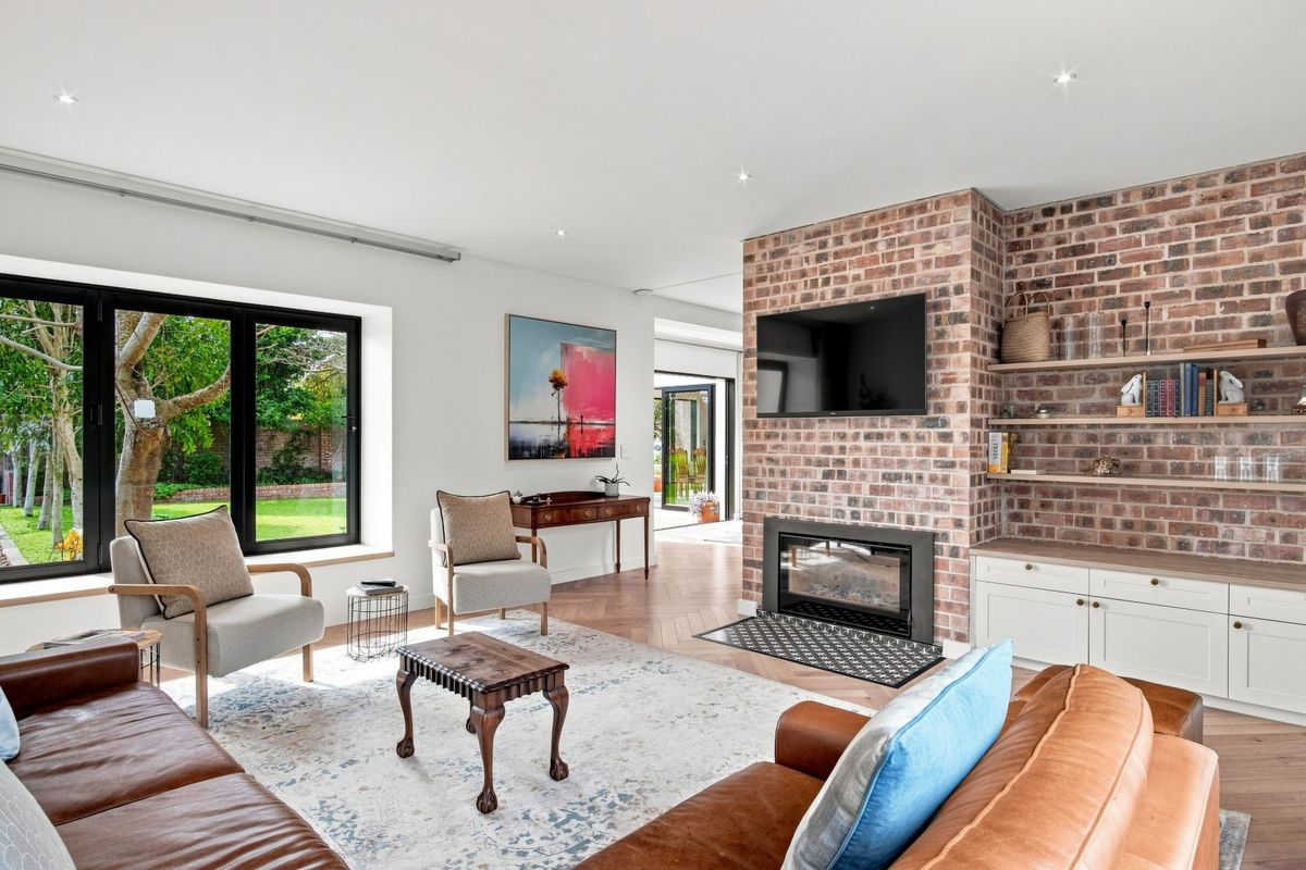 A bright, modern living room featuring an exposed brick fireplace, built-in cabinetry with shelving, and herringbone timber flooring with large windows overlooking the garden.
