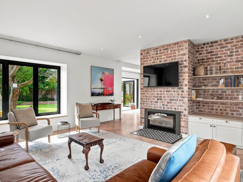 A bright, modern living room featuring an exposed brick fireplace, built-in cabinetry with shelving, and herringbone timber flooring with large windows overlooking the garden.