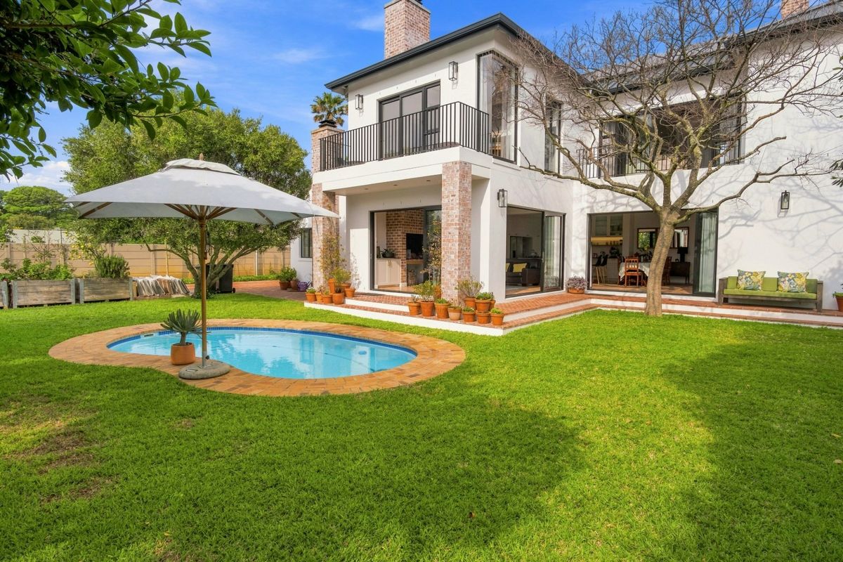 Rear view of a two-story house featuring a landscaped garden, swimming pool, brick patio, and an upper-level balcony with black railings.